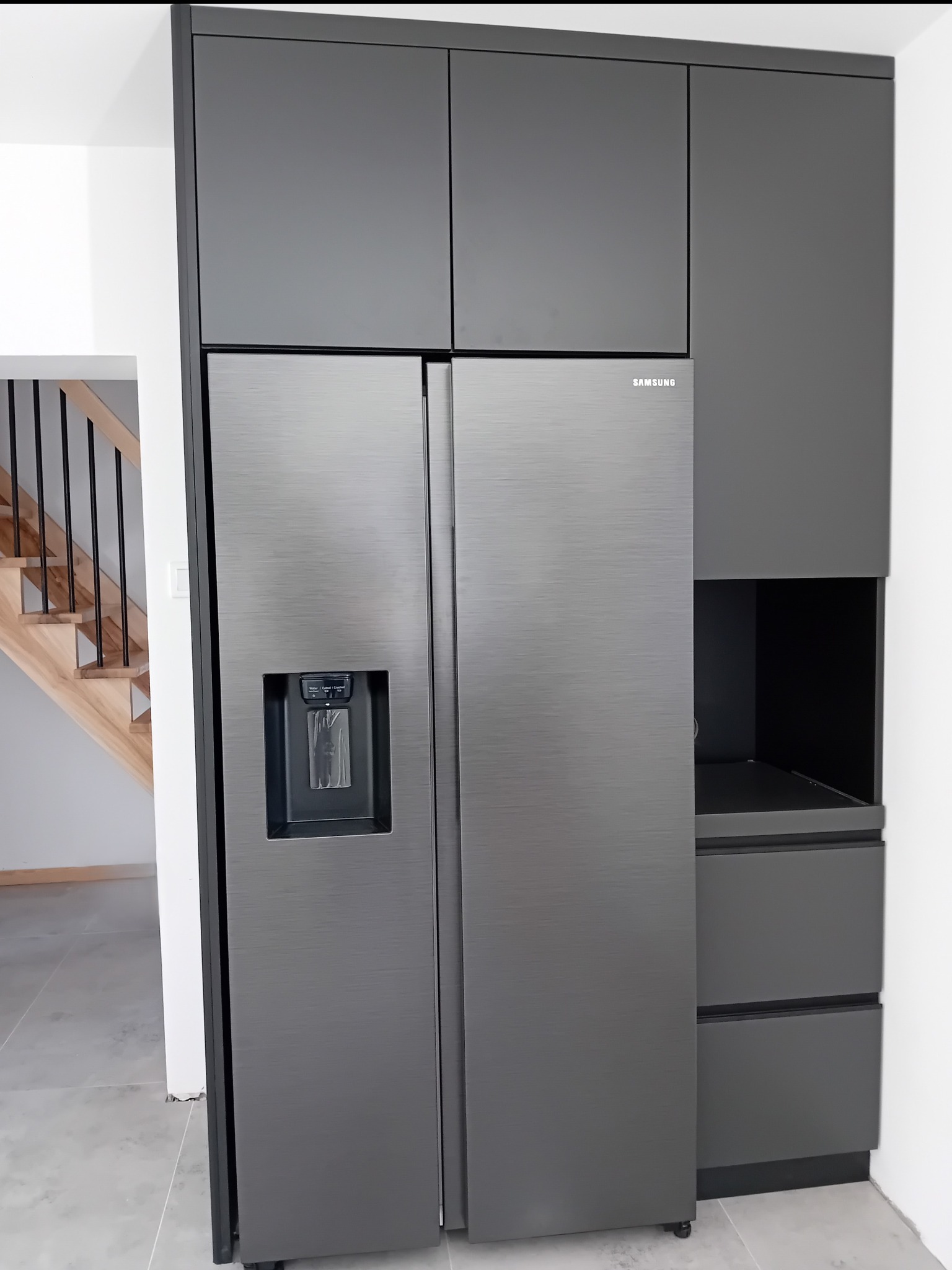 Nowoczesna kuchnia z zabudowaną lodówką Samsung w szarych szafkach, z widocznymi drewnianymi schodami w tle. Minimalistyczny design, proste linie.