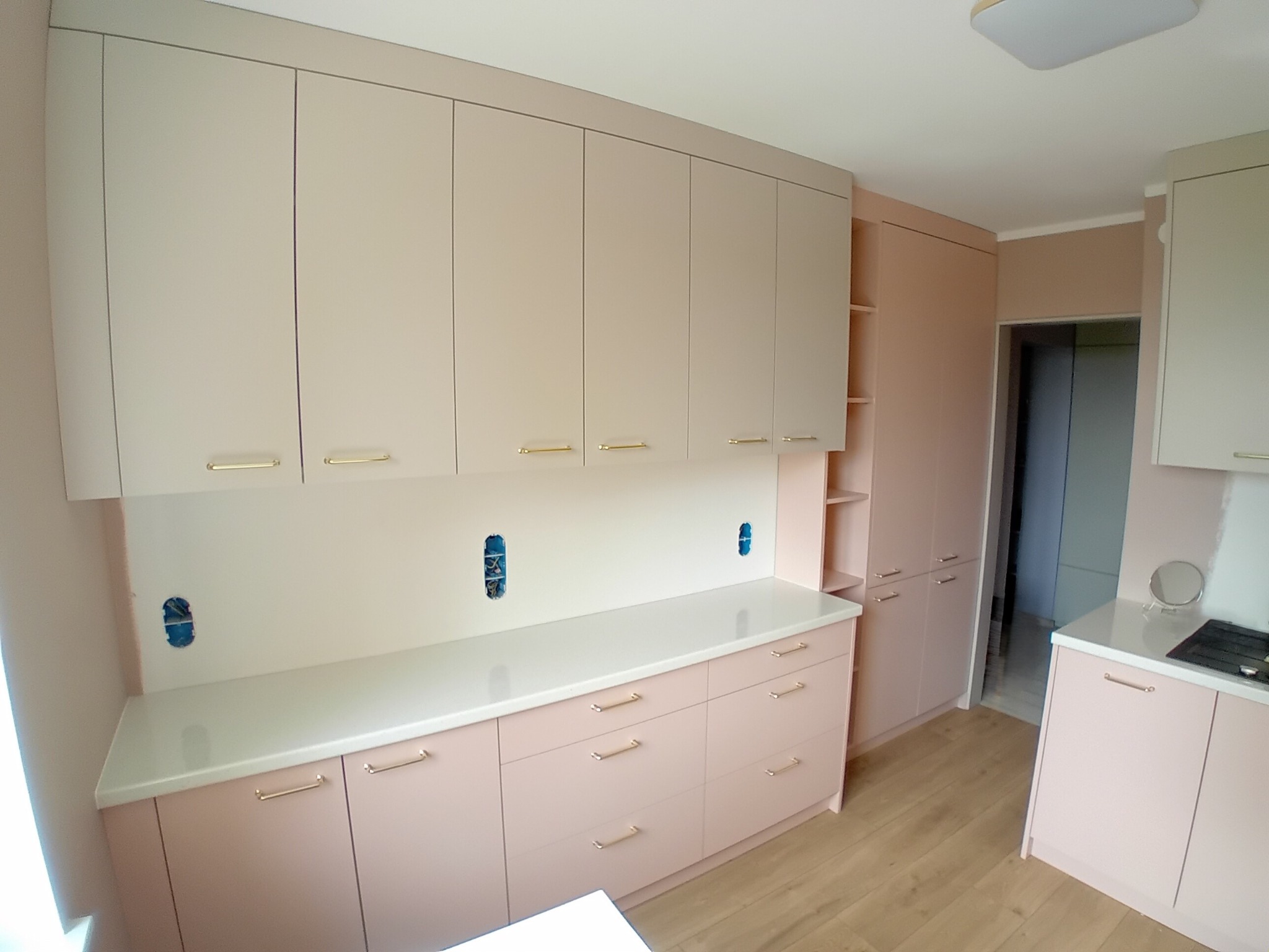 Jasna kuchnia na wymiar z beżowymi frontami i złotymi uchwytami. Widoczne gniazdka elektryczne przed montażem osprzętu, drewniana podłoga i oświetlenie sufitowe.