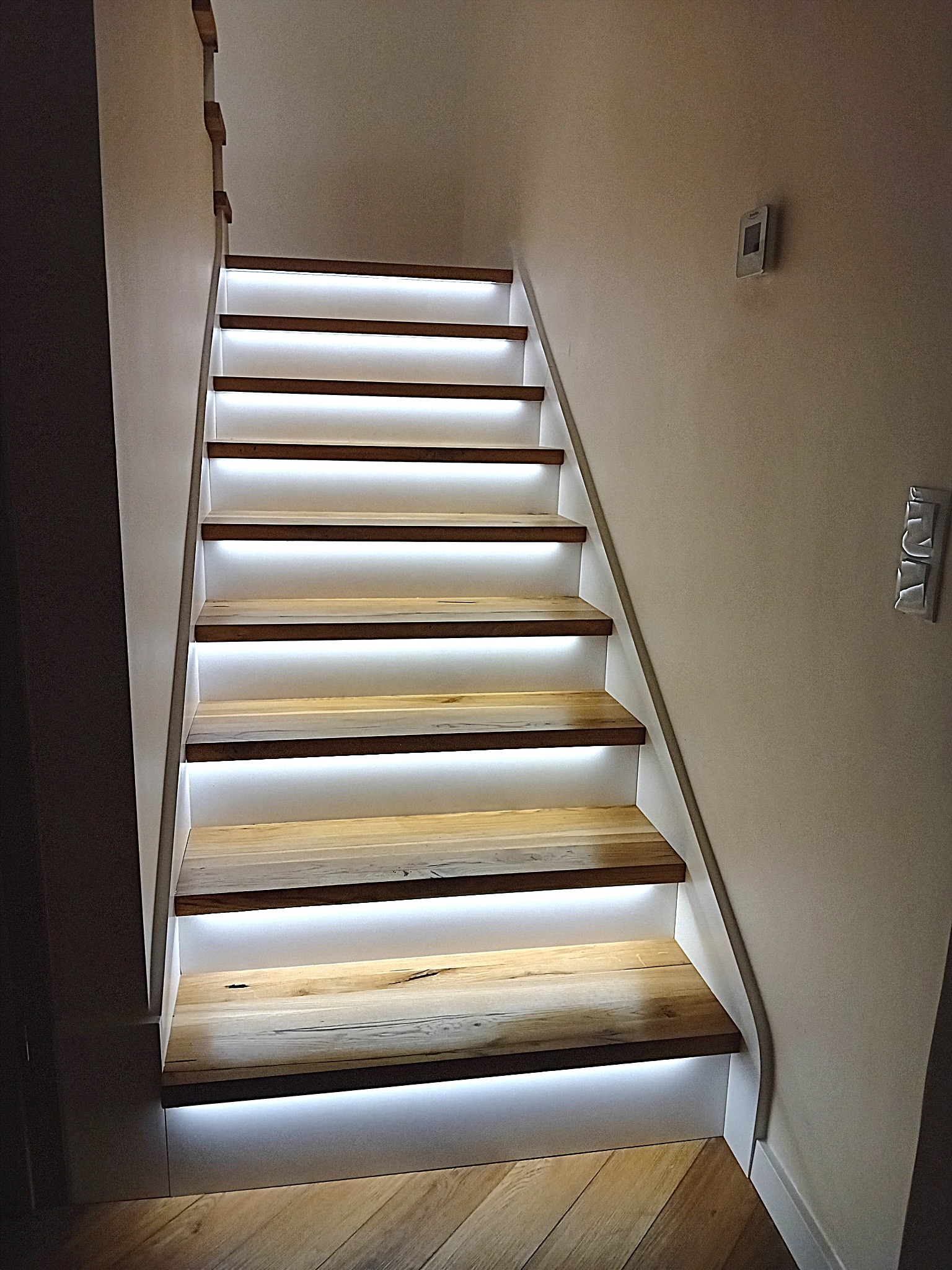 Drewniane schody z podświetleniem LED, widok z dołu. Naturalne drewno i białe elementy. Na ścianie włącznik światła. Parkiet na podłodze.