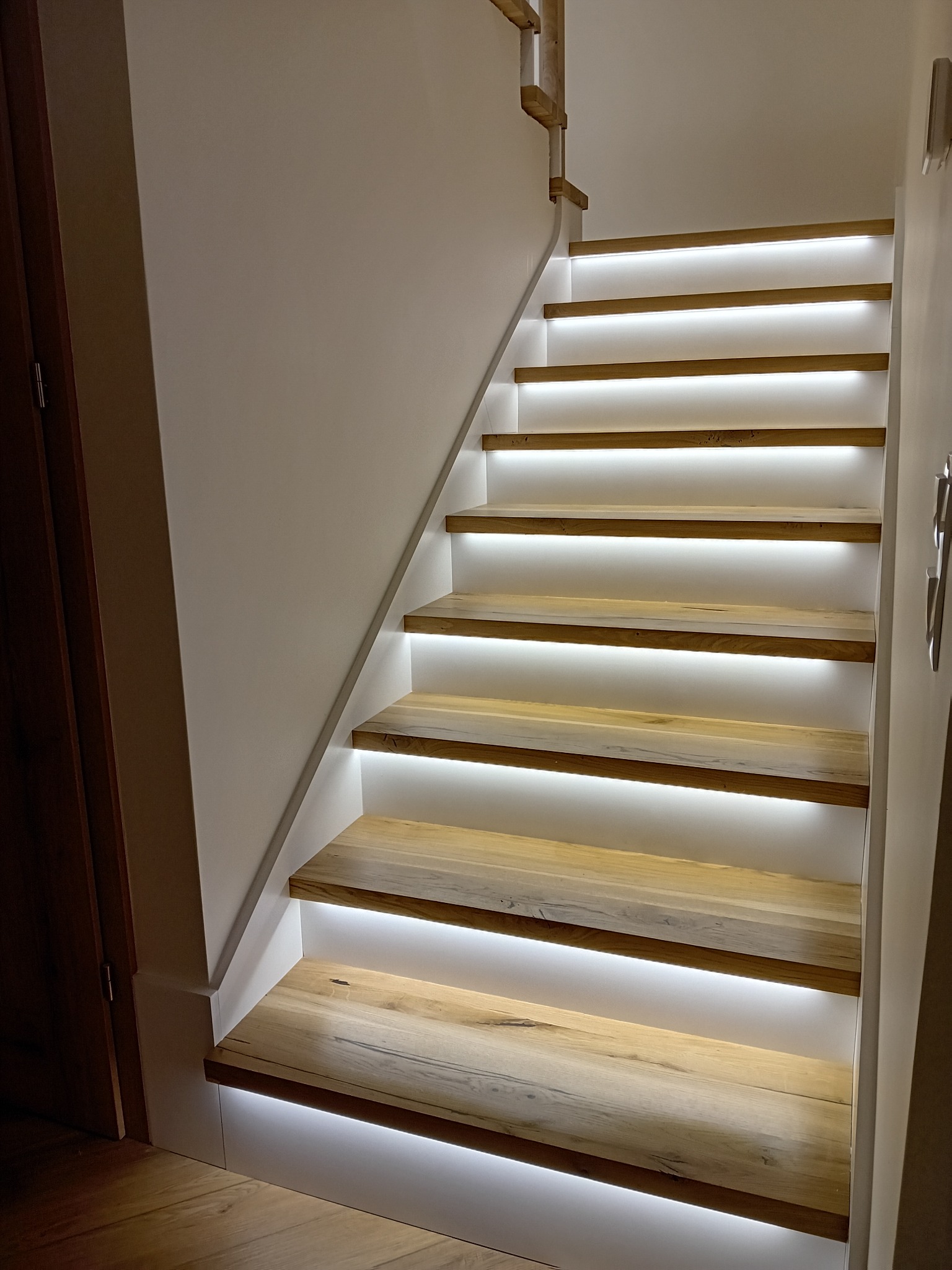 Drewniane schody z subtelnym, białym podświetleniem LED, widok z dołu. Naturalne drewno stopni kontrastuje z bielą ścian i podświetlenia.