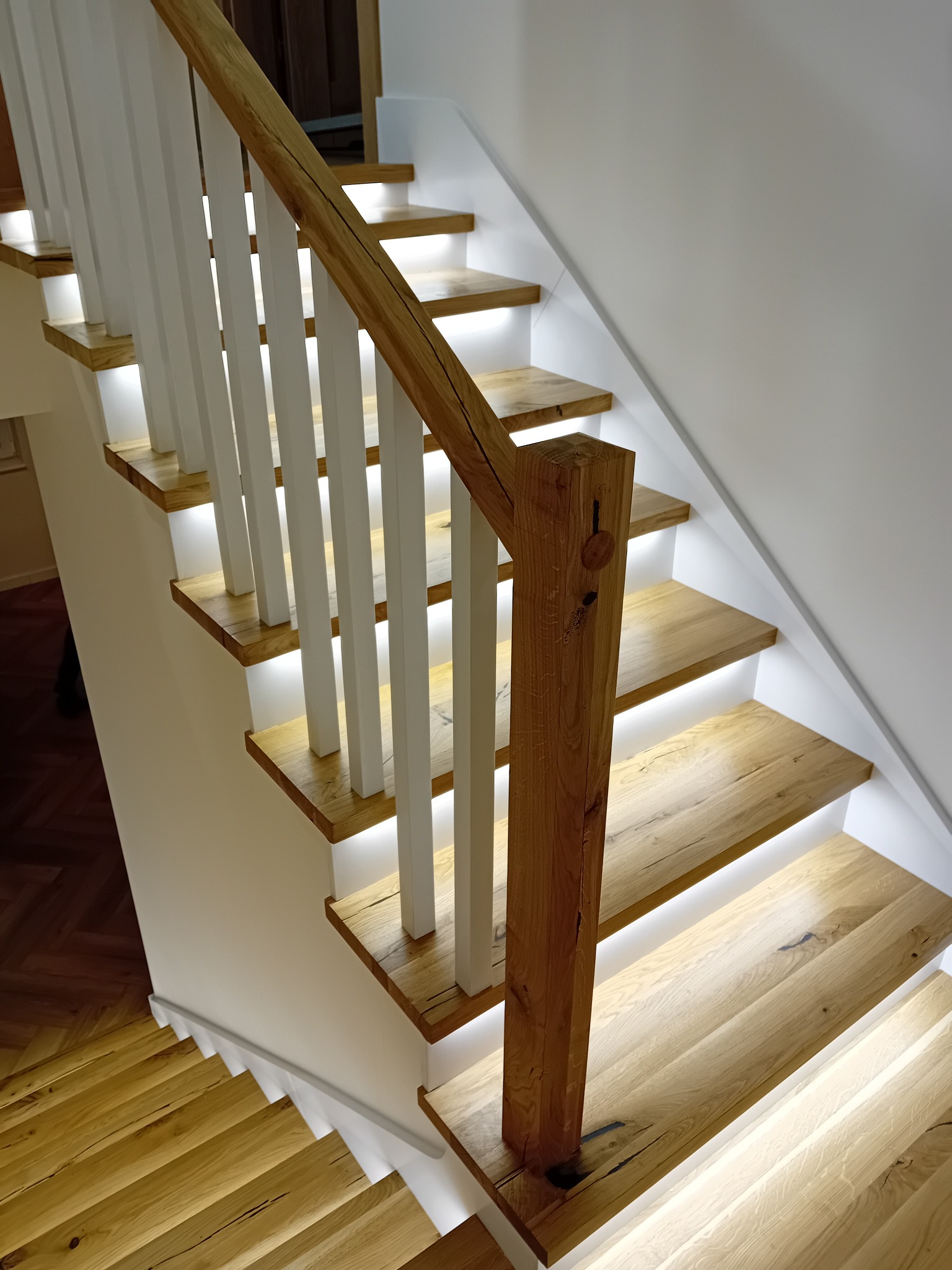 Drewniane schody z białymi balustradami i subtelnym podświetleniem LED, widok z góry, ukazujący naturalną teksturę drewna i nowoczesny design.