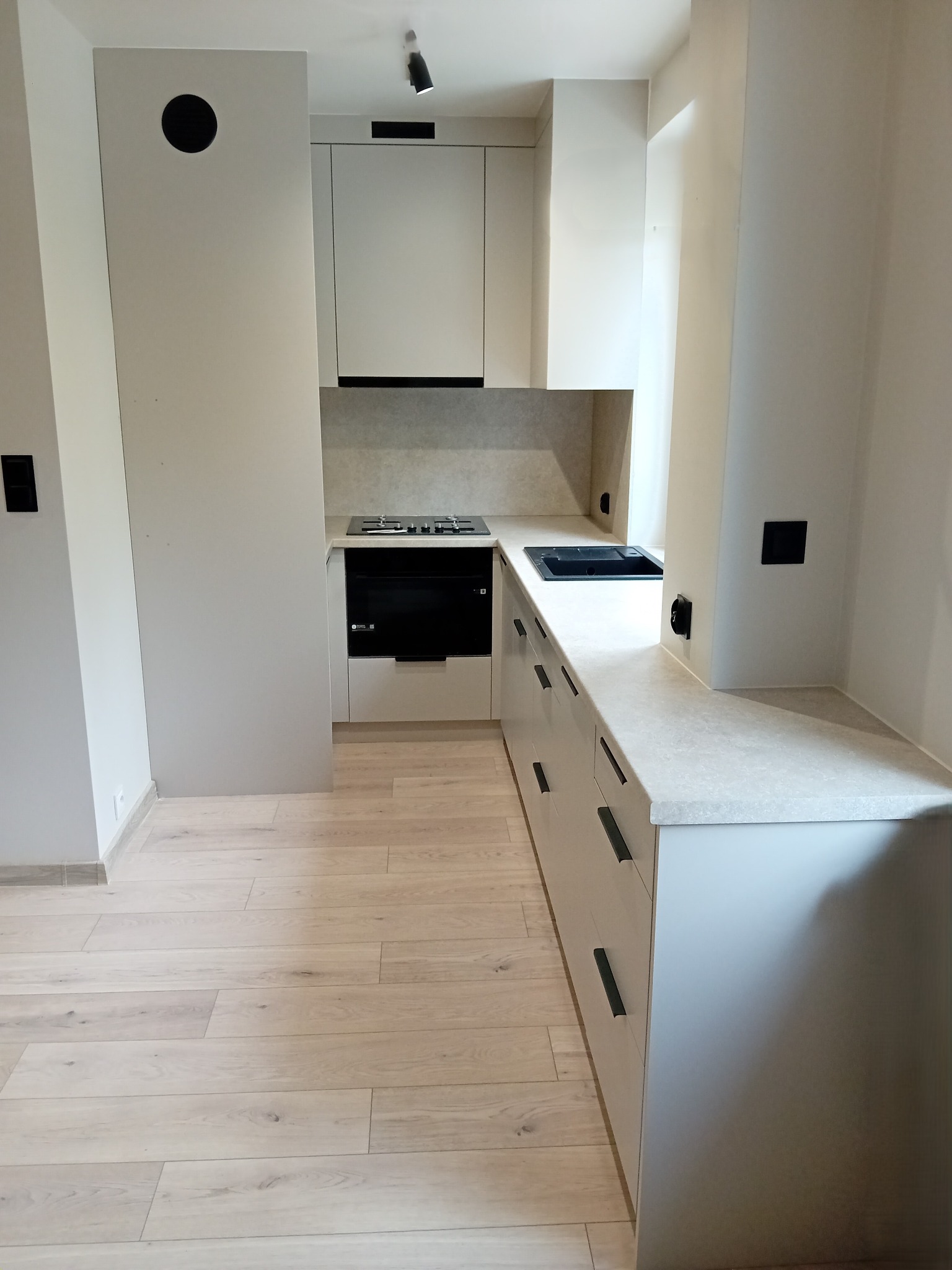 Jasna kuchnia na wymiar z drewnianą podłogą. Ułożenie w kształcie litery L, czarne uchwyty szafek, płyta grzewcza i zlew. Minimalistyczny design.