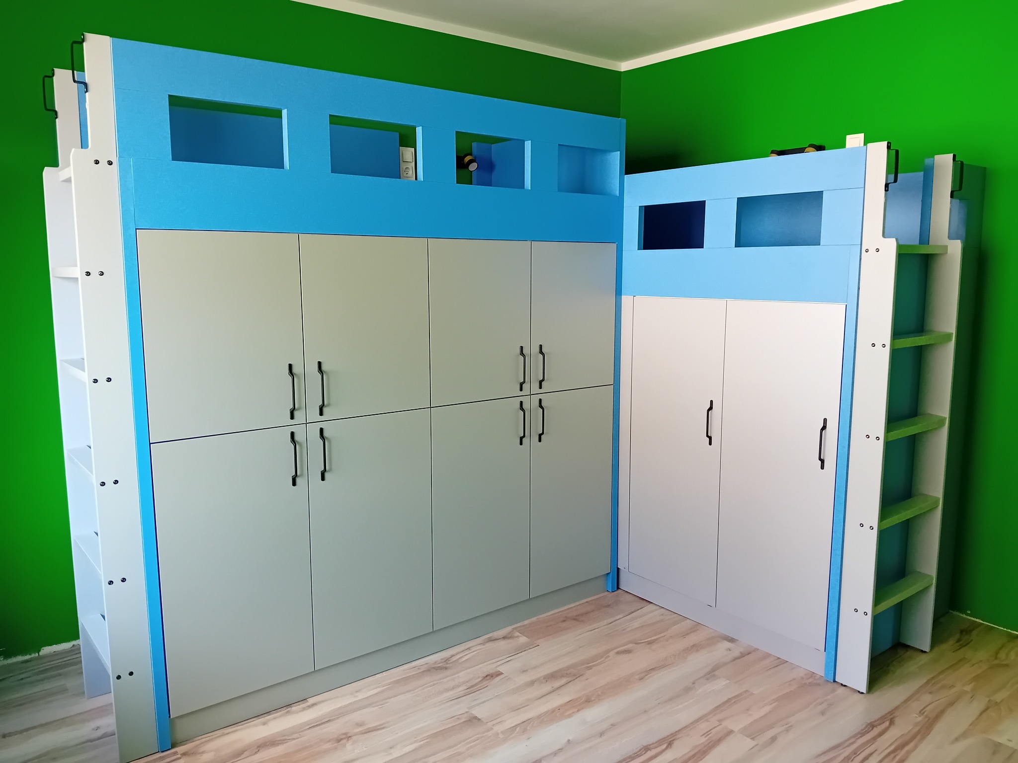 Nietypowe łóżko piętrowe z szafami na wymiar w Goniądzu. Niebieska góra z wnękami i biało-szare szafki z czarnymi uchwytami. Drabinka z boku.