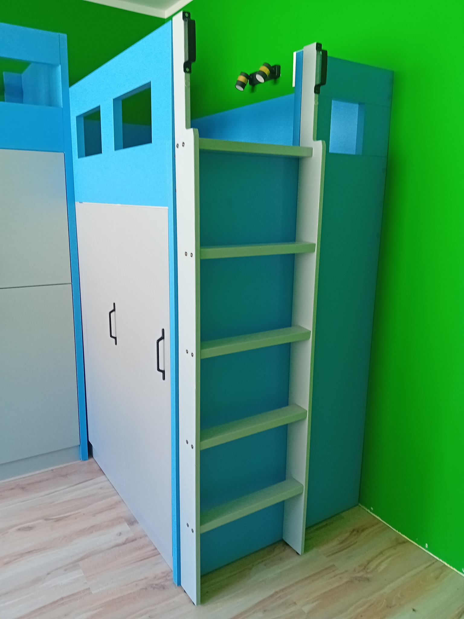 Niebiesko-białe łóżko piętrowe z drabinką i szafą wbudowaną w dolną część, na tle zielonej ściany. Widoczne drewniane panele podłogowe.