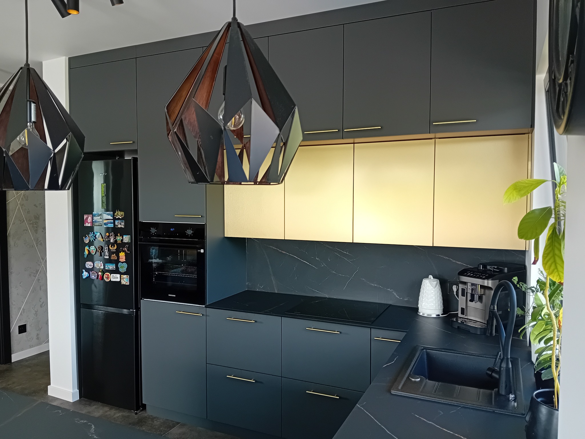 Nowoczesna, grafitowa kuchnia na wymiar z złotymi akcentami, designerskimi lampami i marmurowym blatem. Widok na zabudowę z lodówką i piekarnikiem.