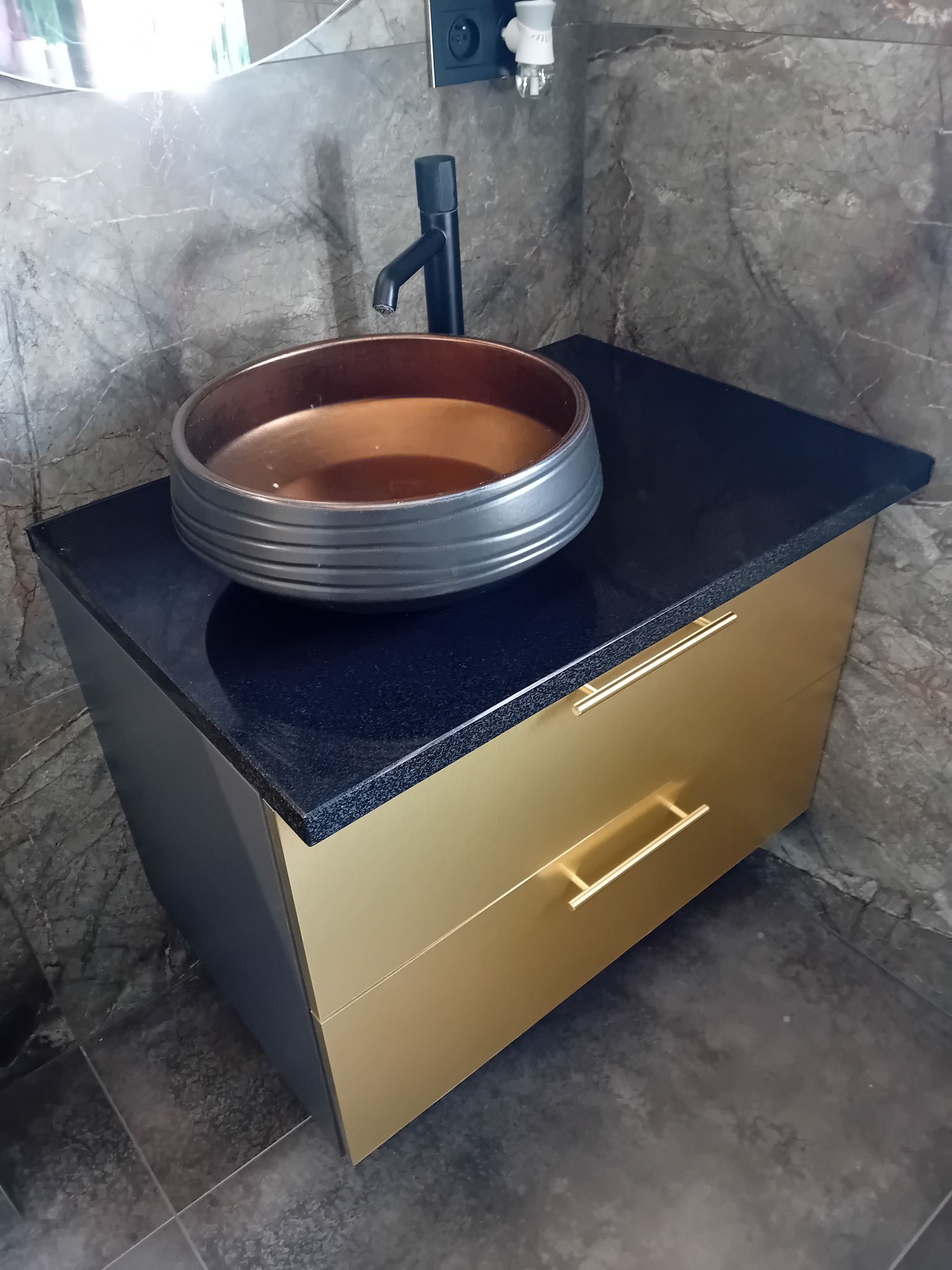 Nowoczesna umywalka nablatowa w odcieniach brązu i szarości, osadzona na czarnym blacie z szafką w kolorze złota. Marmurowe wykończenie ściany i podłogi.