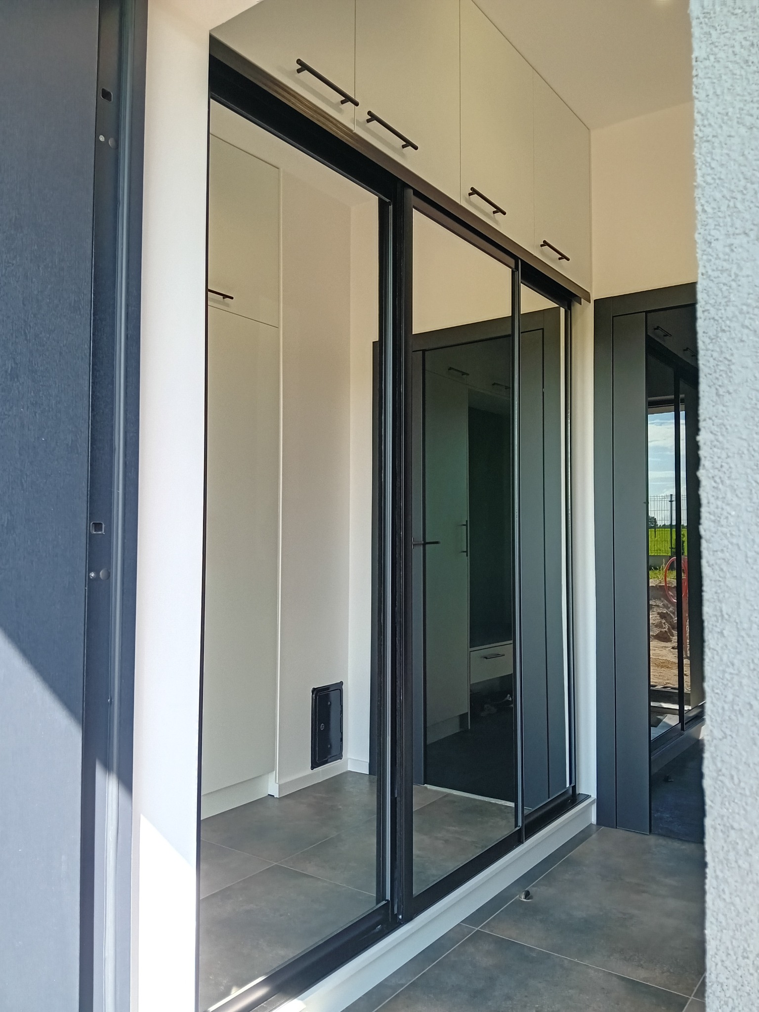 Nowoczesna szafa wnękowa z przesuwnymi lustrzanymi drzwiami w czarnej ramie, z górnymi szafkami w jasnym odcieniu, widoczna z perspektywy wnętrza.