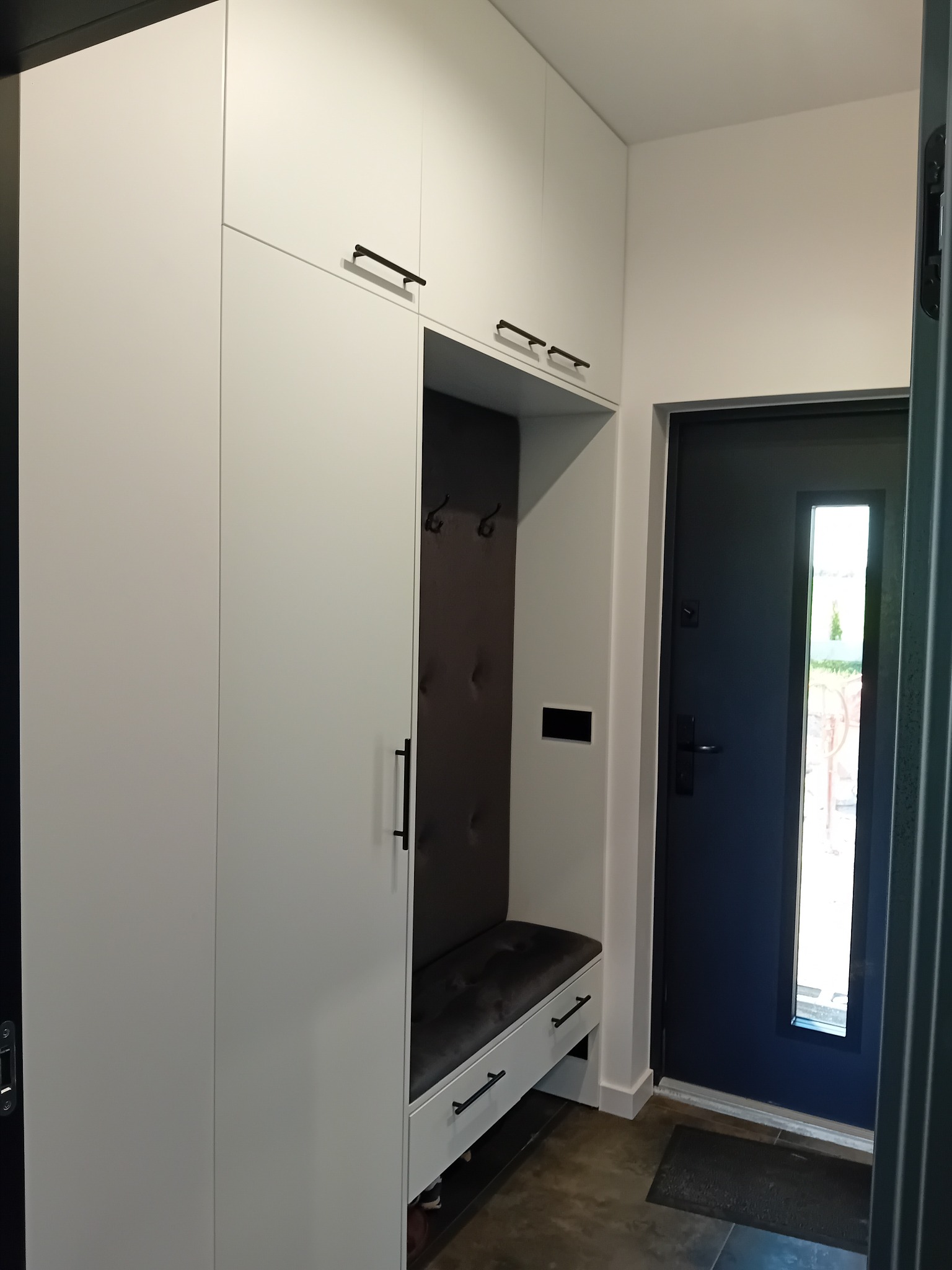 Biała szafa wnękowa z siedziskiem i wieszakami, czarne uchwyty, minimalistyczny design, widok na drzwi wejściowe. Nowoczesne rozwiązanie do przedpokoju.