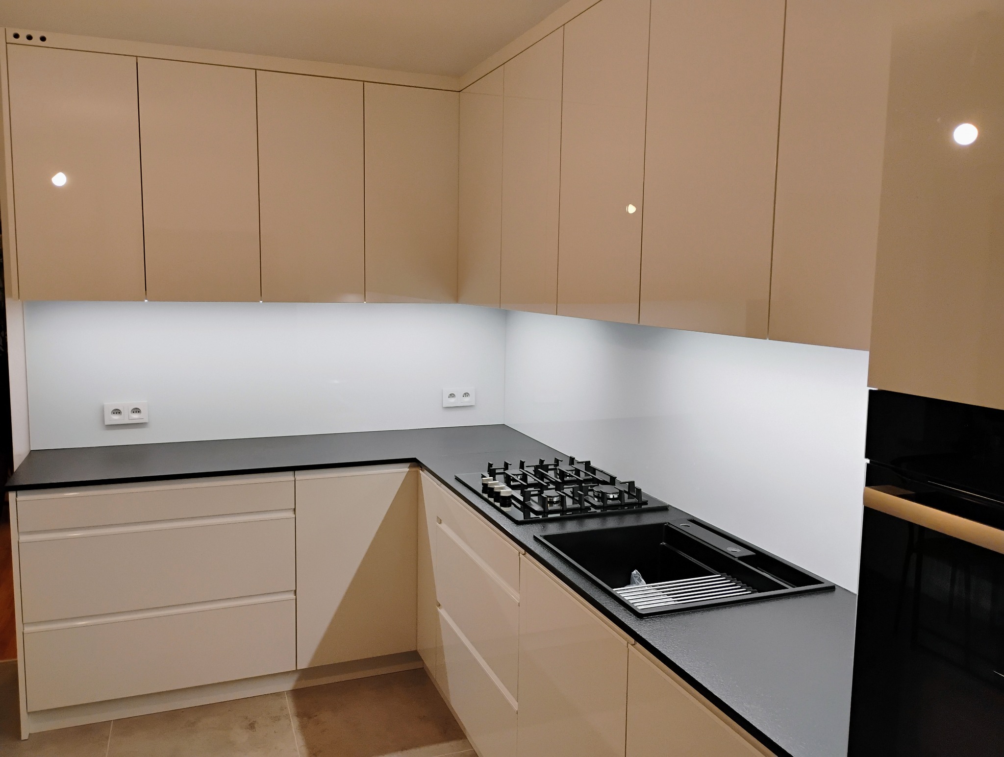 Beżowa kuchnia z połyskiem: szafki górne i dolne, czarny blat, płyta gazowa, zlewozmywak. Nowoczesne oświetlenie LED podszafkowe. Gniazdka elektryczne w białej obudowie.