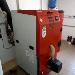 Kocioł na paliwo stałe marki Kottlebahn, z widocznymi elementami instalacji, takimi jak rury i zawory, zainstalowany w kotłowni z jasnymi płytkami na podłodze.