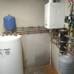 Biały zasobnik ciepłej wody użytkowej Kospel z niebieskim naczyniem przeponowym, wiszący biały piec gazowy, czarny wymiennik ciepła i rozbudowana instalacja hydrauliczna z zaworami kulowymi...