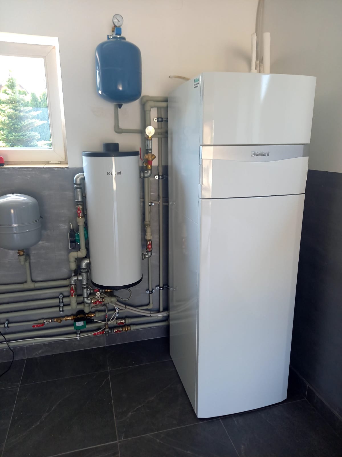 Biały, nowoczesny piec gazowy Vaillant z podłączonymi rurami i zbiornikiem wyrównawczym w kotłowni z oknem. Widoczne liczne zawory i manometry.