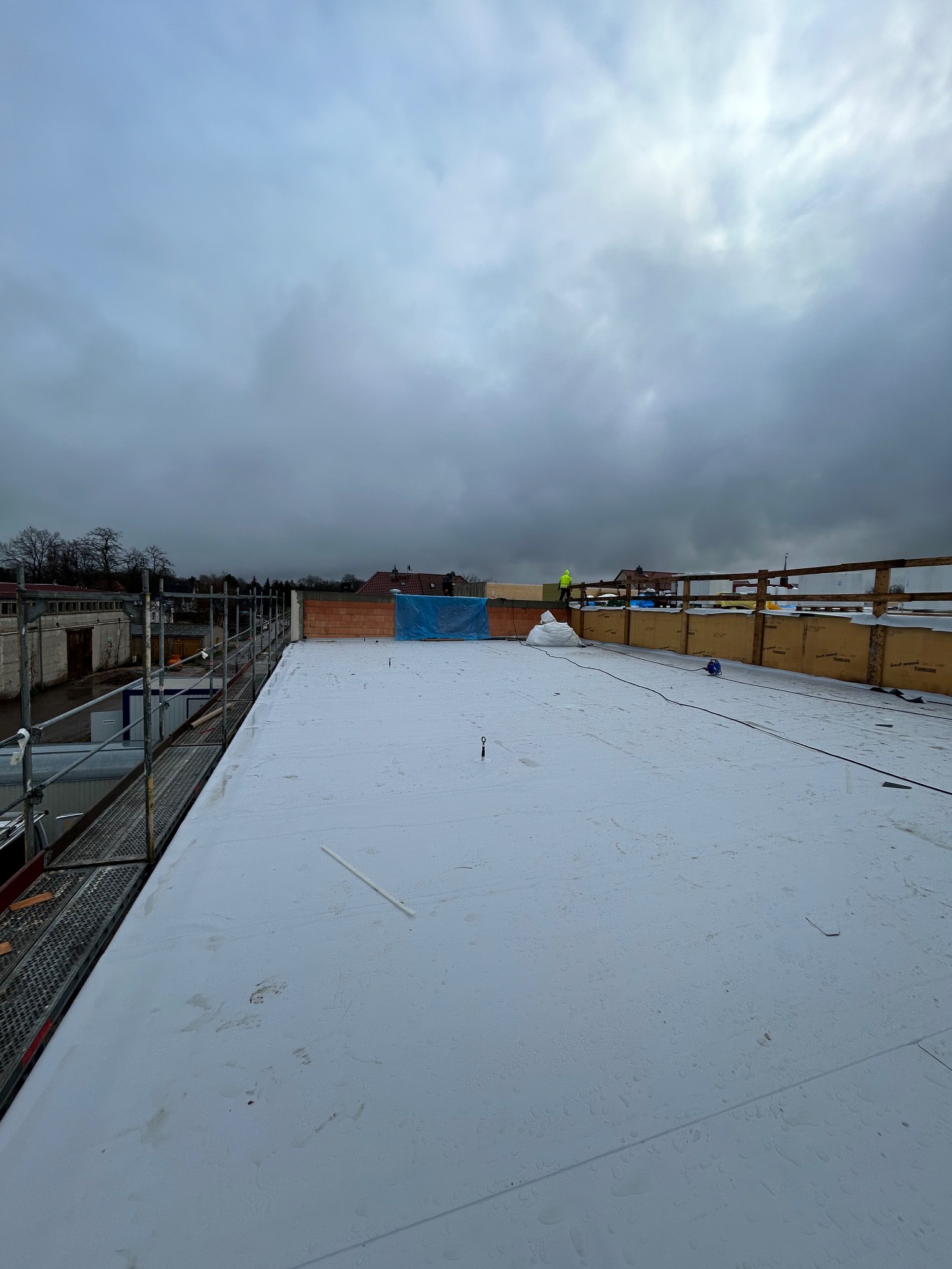 Widok na płaski dach pokryty białą membraną w Szczecinie, z widocznymi śladami zabrudzeń i pracującym dekarzem w oddali, pochmurne niebo w tle.