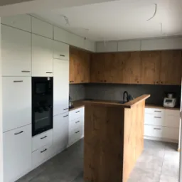 Nowoczesna kuchnia z białymi i drewnianymi szafkami, czarnymi uchwytami i wyspą kuchenną z drewna w odcieniach beżu, szare płytki podłogowe, niedokończone podłączenia elektryczne na suficie.