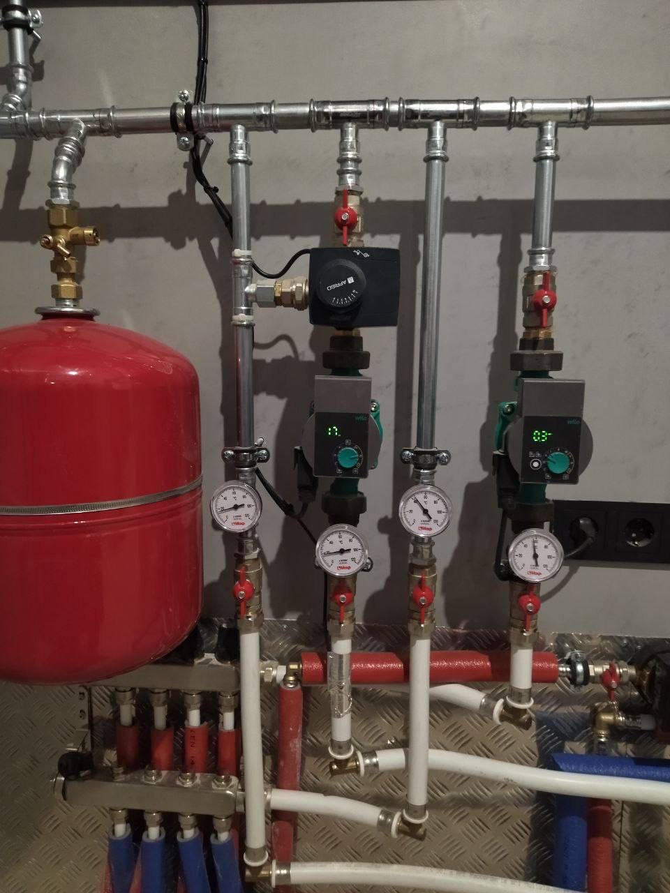Rozbudowana instalacja centralnego ogrzewania z czerwonym zbiornikiem wyrównawczym, manometrami i pompami obiegowymi na tle metalowej ściany z fakturą rombów.