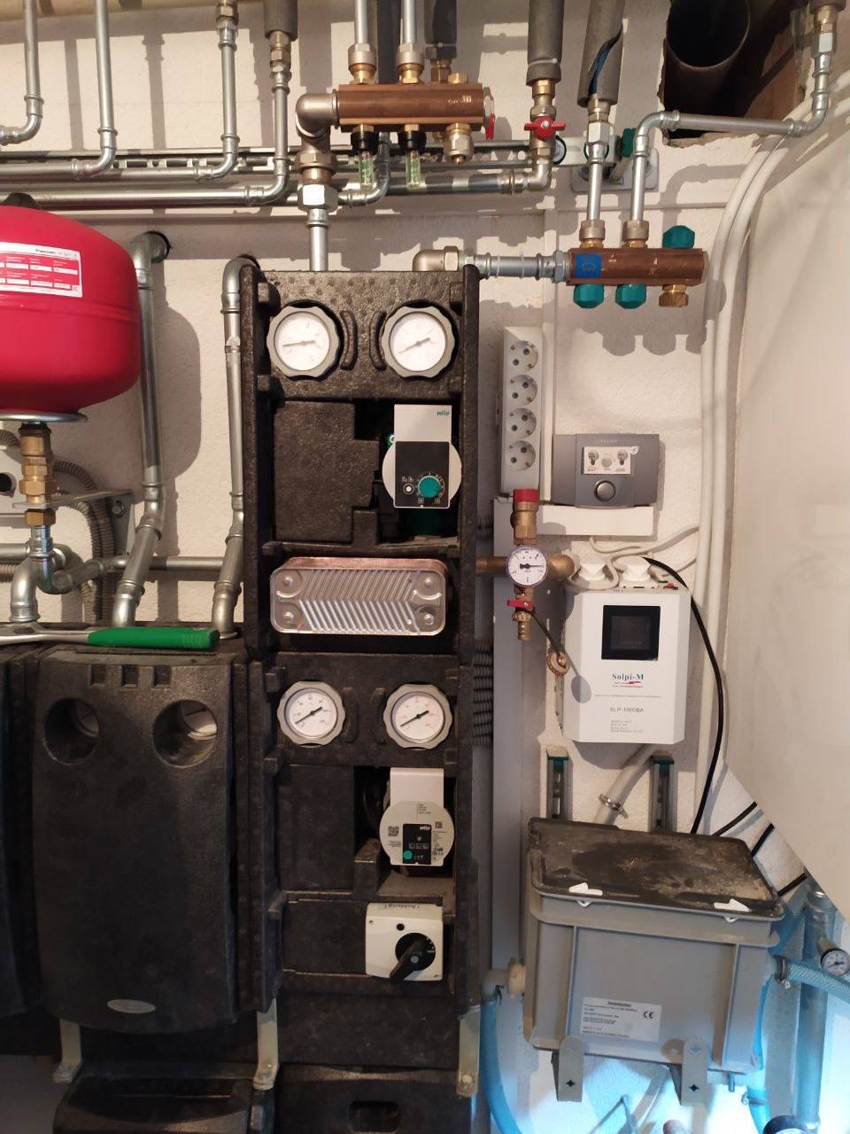 Rozbudowana instalacja grzewcza w kotłowni z czerwonym zbiornikiem wyrównawczym, licznymi rurami, zaworami, manometrami i sterownikami, zamontowana na ścianie wewnątrz pomieszczenia.