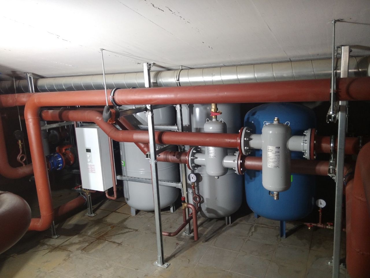 Złożona instalacja centralnego ogrzewania z rurami w kolorze ceglastym, zbiornikami akumulacyjnymi i białym kotłem wiszącym w kotłowni.