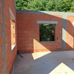 Wnętrze budynku w trakcie budowy, widoczne ściany z czerwonej cegły z otworami okiennymi, częściowo zabetonowane nadproża, niebieski wąż na podłodze, cień od słońca na posadzce.