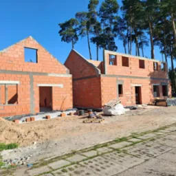 Dwa budynki w stanie surowym otwartym z czerwonej cegły, z widocznymi otworami okiennymi i drzwiowymi, na tle lasu sosnowego i błękitnego nieba, z fragmentem niebieskiego samochodu dostawczego po...