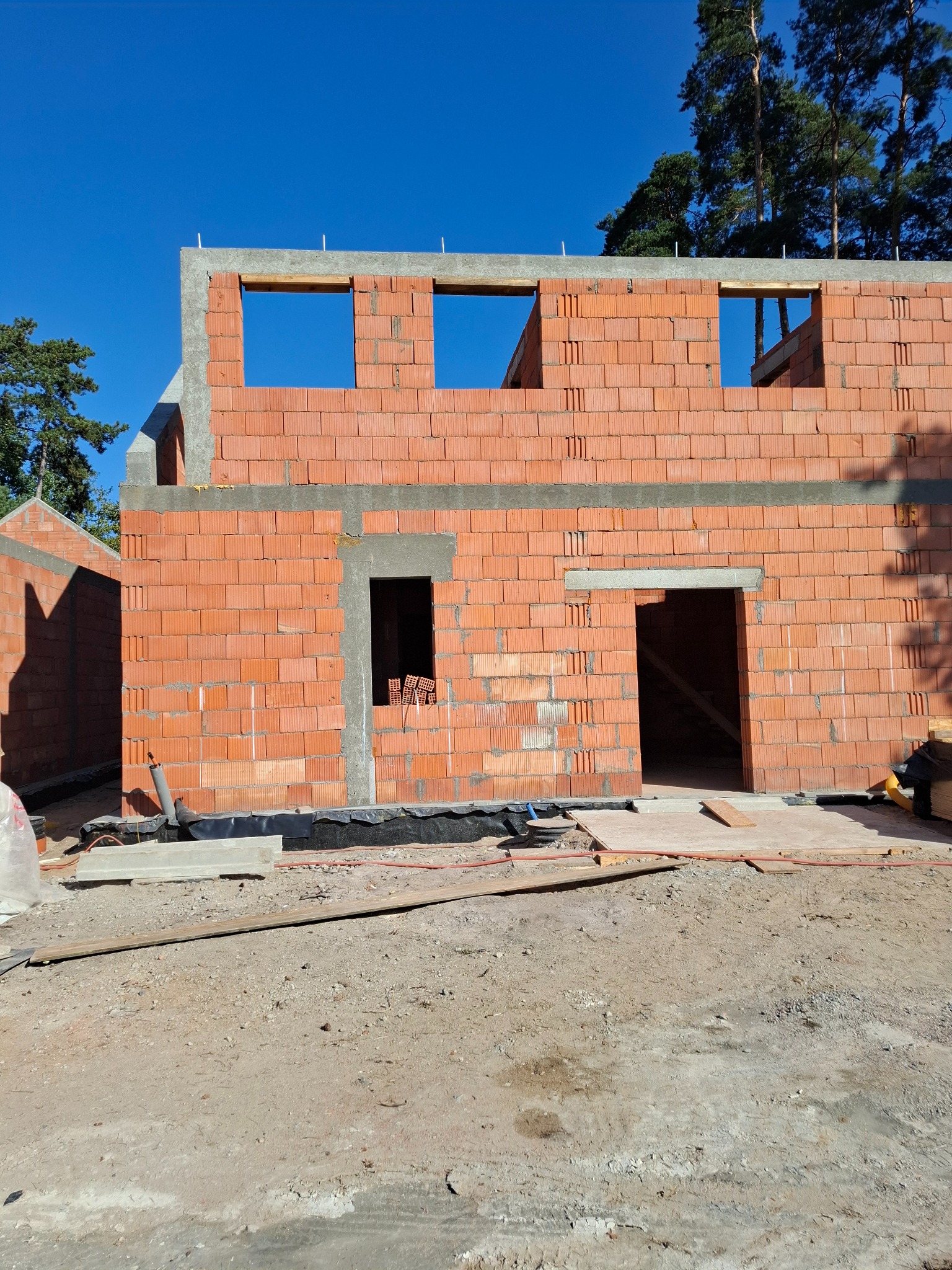 Surowa bryła budynku w trakcie wznoszenia z czerwonej cegły kratówki, widoczne otwory okienne i drzwiowe, betonowe nadproża i fundamenty, otoczenie placu budowy z piaskiem i drewnem.