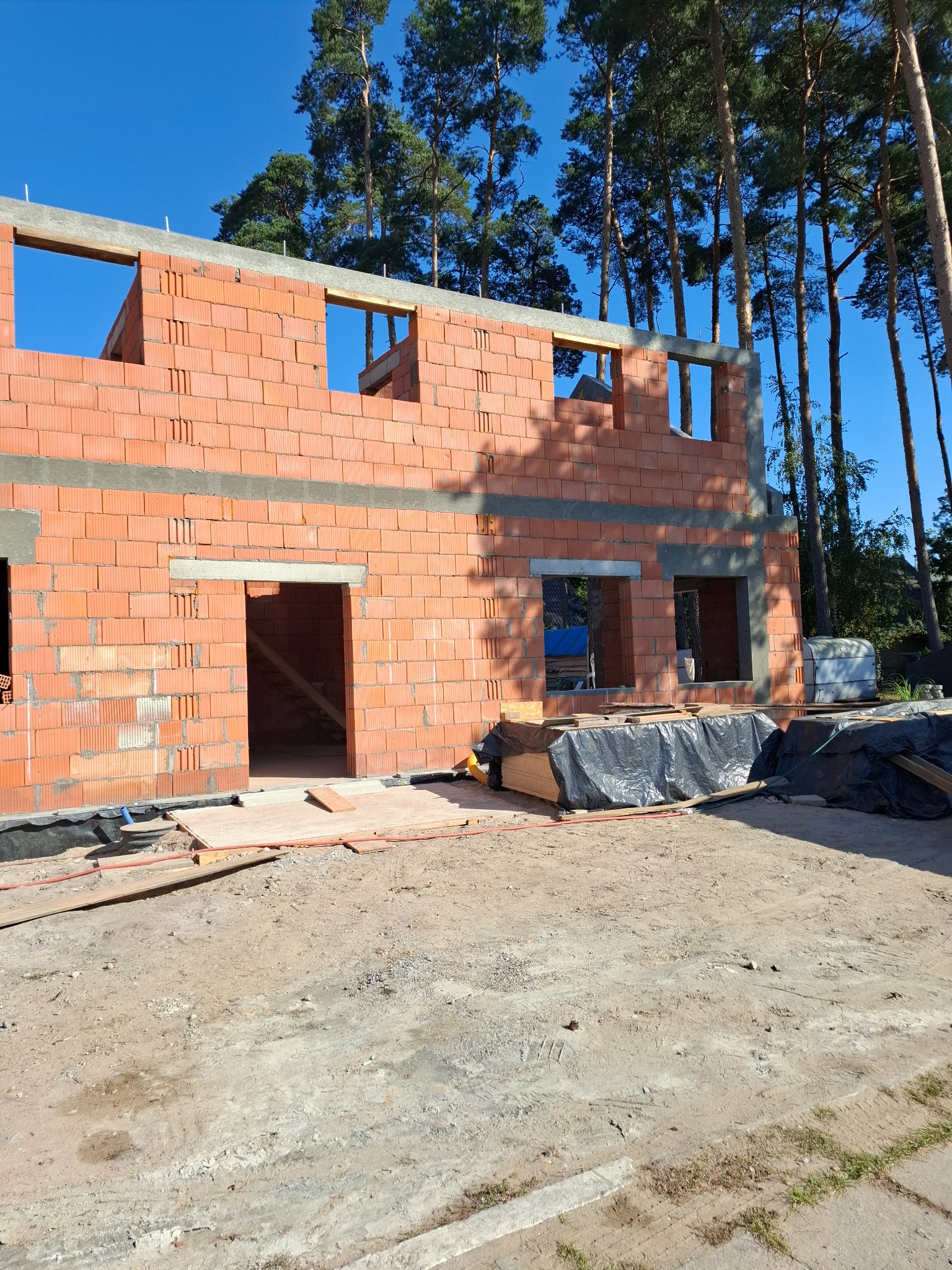 Wznoszenie ścian z czerwonej cegły ceramicznej na budowie domu jednorodzinnego, widoczne otwory okienne i drzwiowe, w tle wysokie sosny i błękitne niebo.
