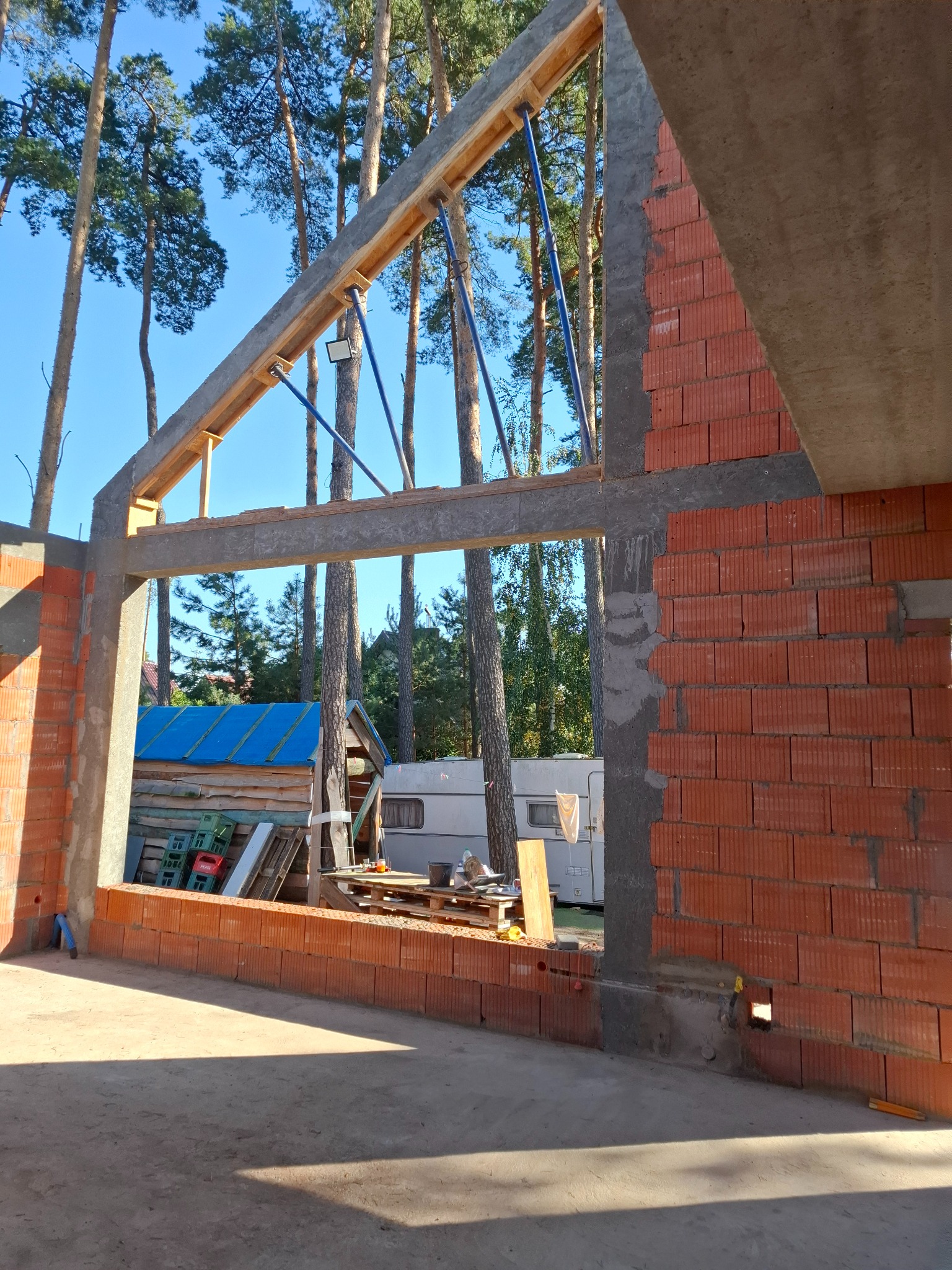 Wznoszenie ściany z czerwonej cegły dziurawki z widocznym szalunkiem i podporami, w tle las i przyczepa kempingowa.