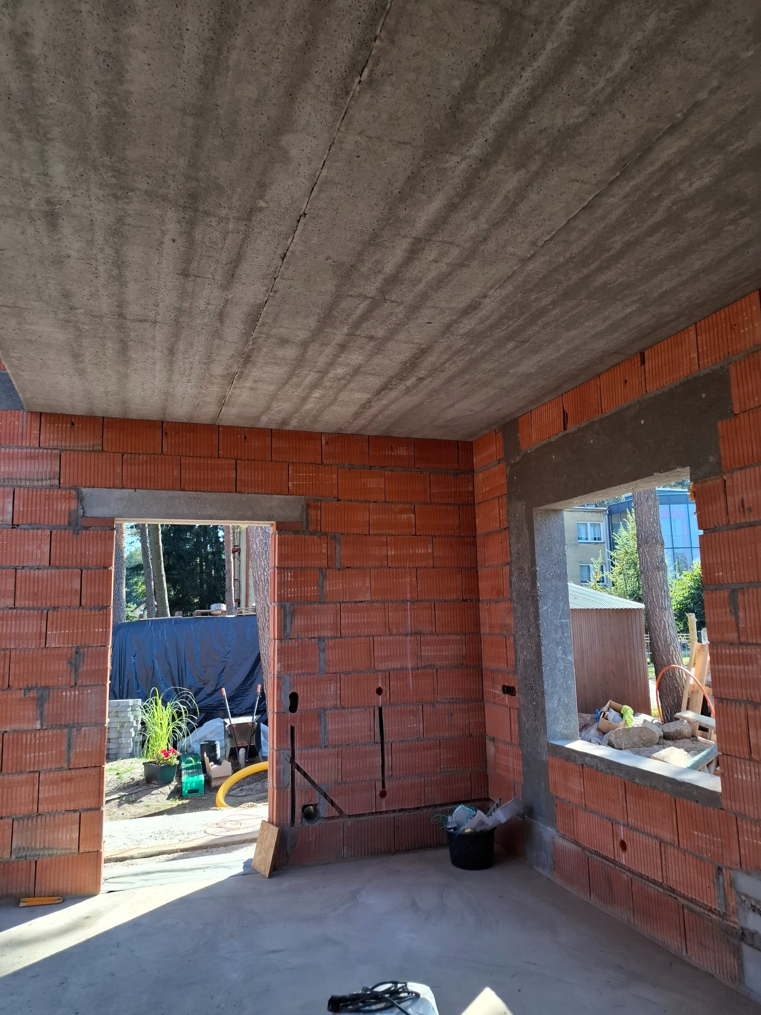 Surowy stan wnętrza domu w trakcie budowy: ściany z czerwonej cegły, betonowy strop, otwory okienne i drzwiowe, widoczne instalacje, narzędzia budowlane w tle.