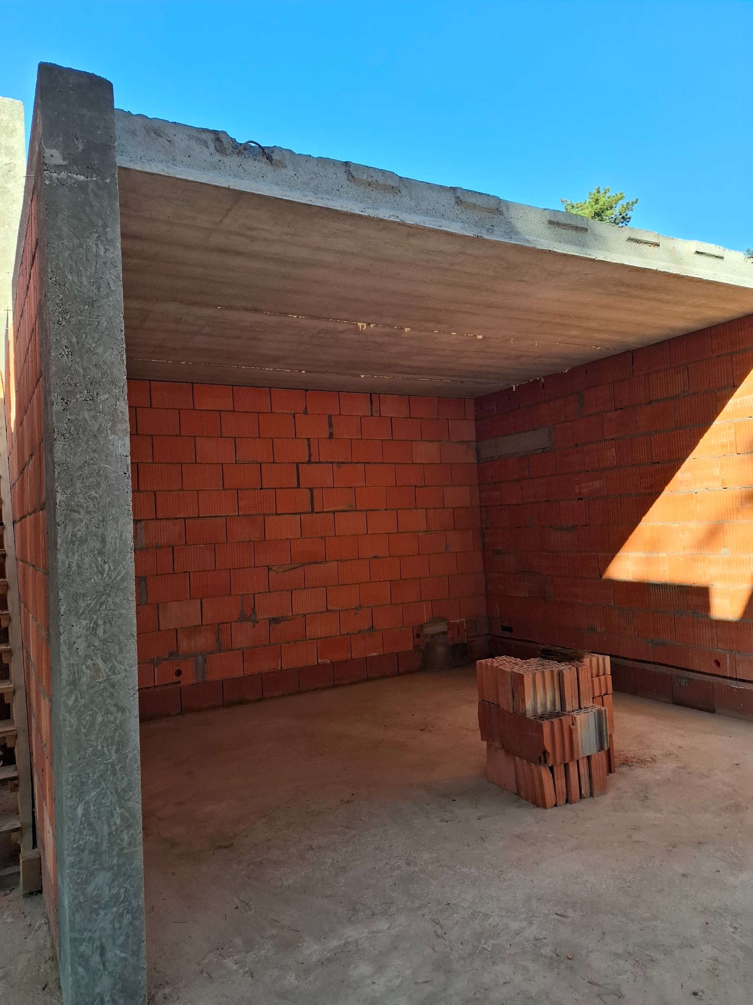 Surowy stan budynku w trakcie budowy, widoczne ściany z czerwonej cegły, betonowy strop i filar, na podłodze ułożony stos cegieł.