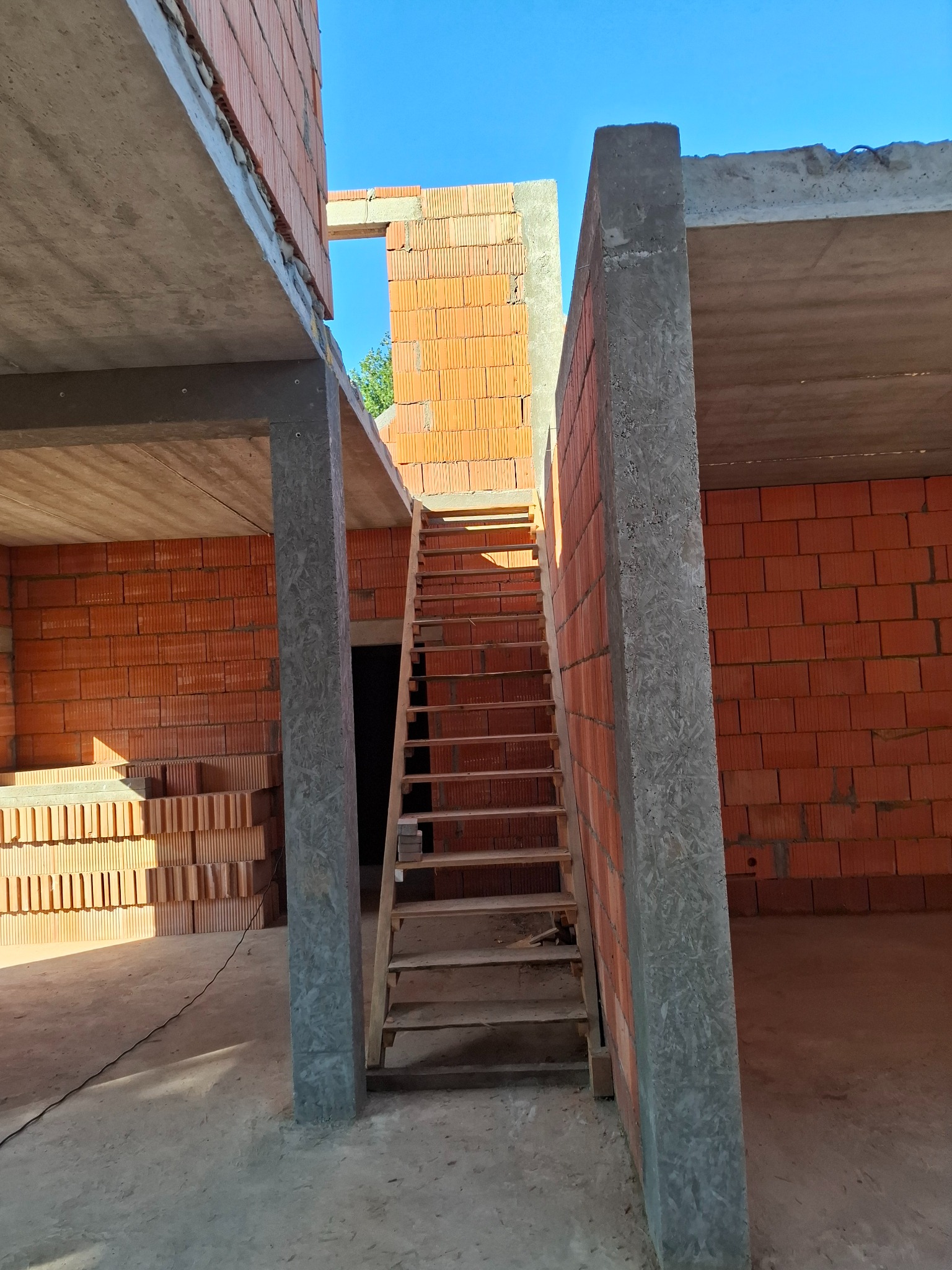 Wnętrze surowego domu w trakcie budowy: ściany z czerwonej cegły, betonowe stropy i słupy, drewniane schody tymczasowe.