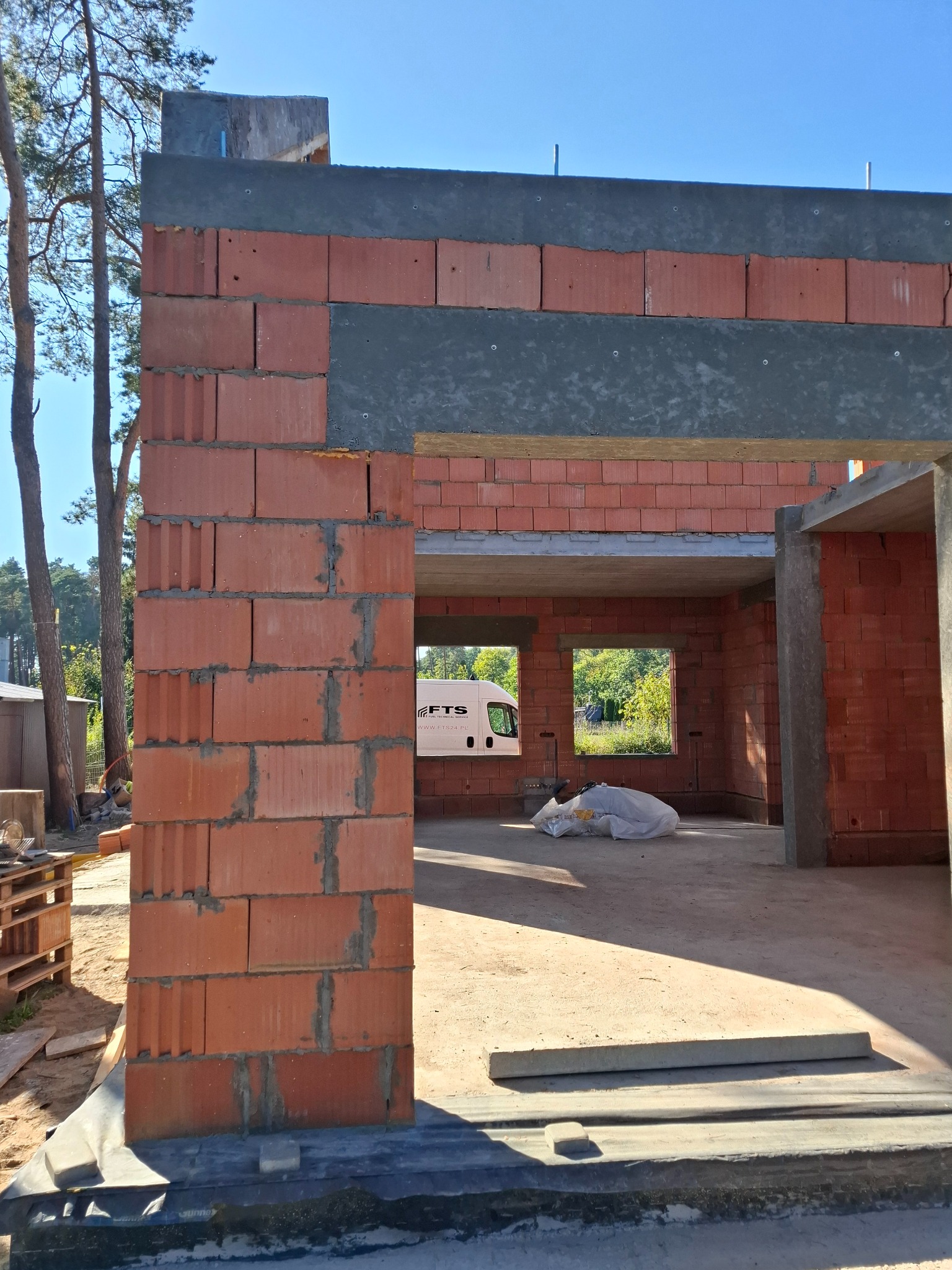 Surowy stan domu murowanego z czerwonej cegły, widoczne nadproża, z zewnątrz widok na las i zaparkowany biały samochód dostawczy, wewnątrz worki z materiałami budowlanymi.