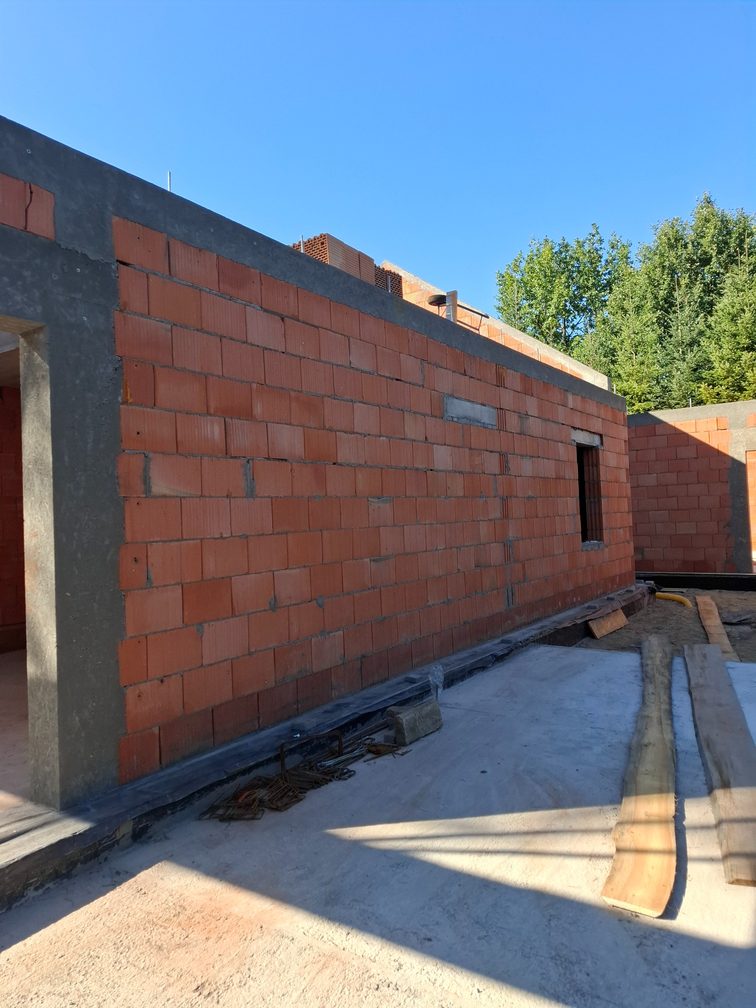 Wznoszenie ścian parterowego budynku z czerwonej cegły ceramicznej, widoczne otwory okienne, szary wieniec żelbetowy, błękitne niebo i zieleń drzew w tle.