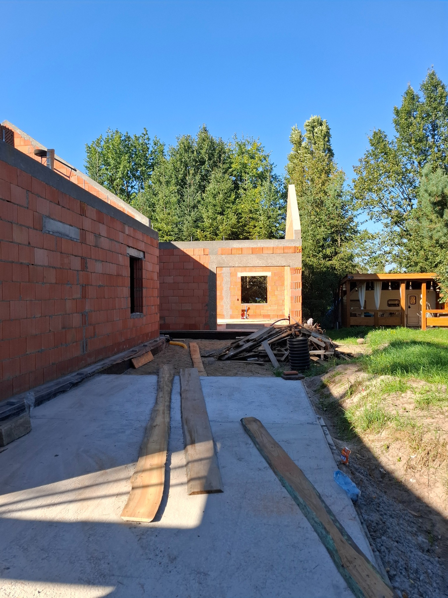 Budowa domu jednorodzinnego w Stargardzie w trakcie wznoszenia ścian z czerwonej cegły, widoczne fundamenty i częściowo postawione ściany, w tle zieleń drzew i altana.