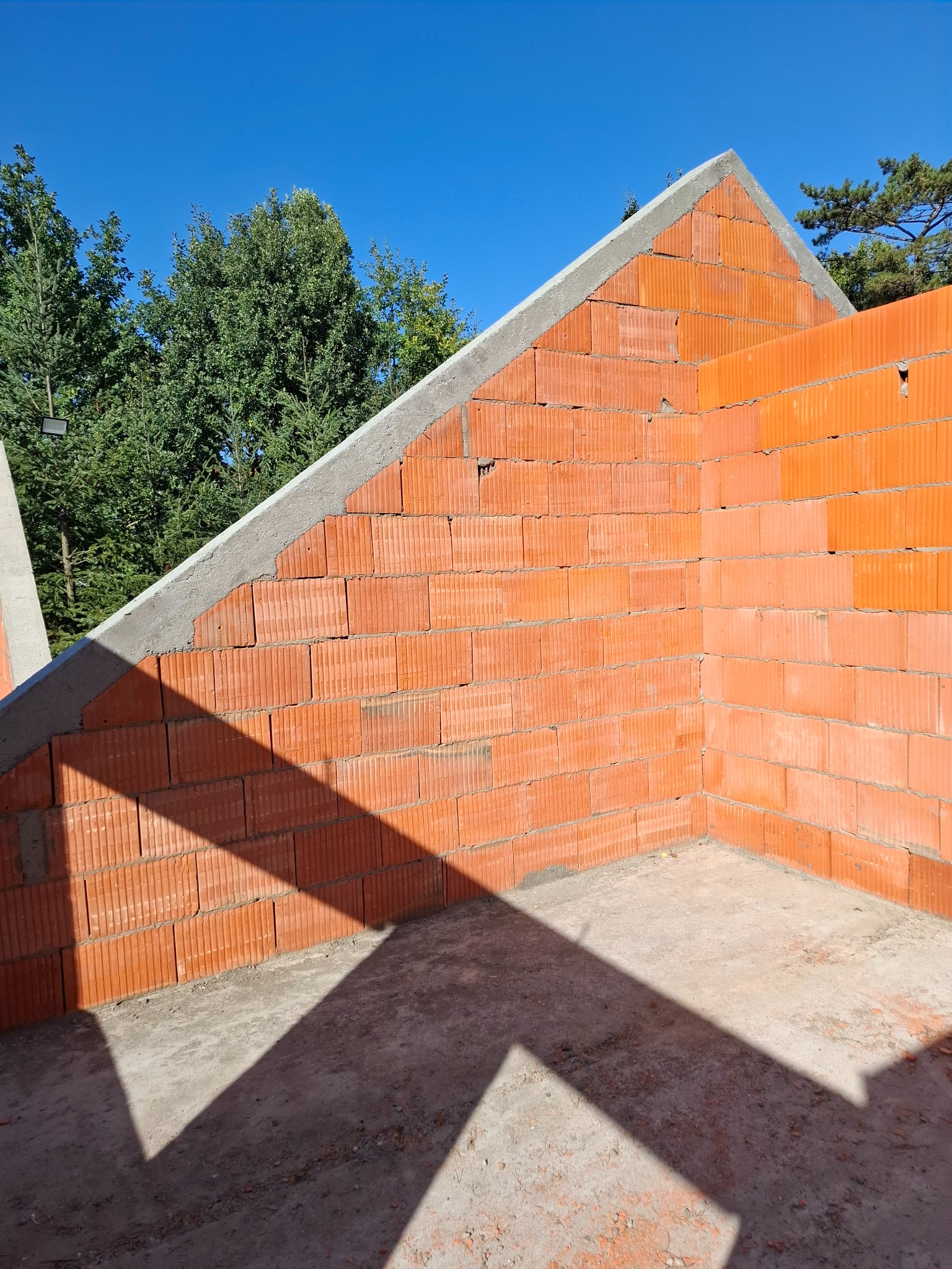 Wewnętrzny narożnik budynku w surowym stanie, wzniesiony z czerwonych bloczków ceramicznych, zakończony szarym wieńcem, z widocznym cieniem konstrukcji dachu na podłodze.