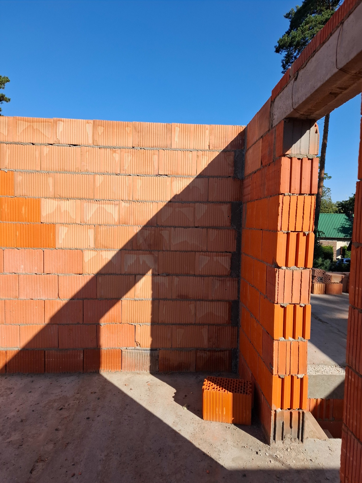 Wznoszone ściany z czerwonej ceramiki poryzowanej, widoczny otwór drzwiowy z nadprożem betonowym, cień na murze w słoneczny dzień.