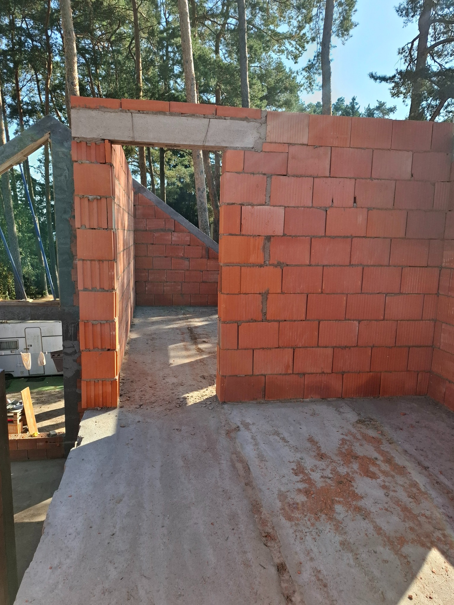 Wznoszenie ścian z czerwonych bloczków ceramicznych na betonowym fundamencie, widoczny otwór drzwiowy z nadprożem, w tle las iglasty i przyczepa kempingowa.