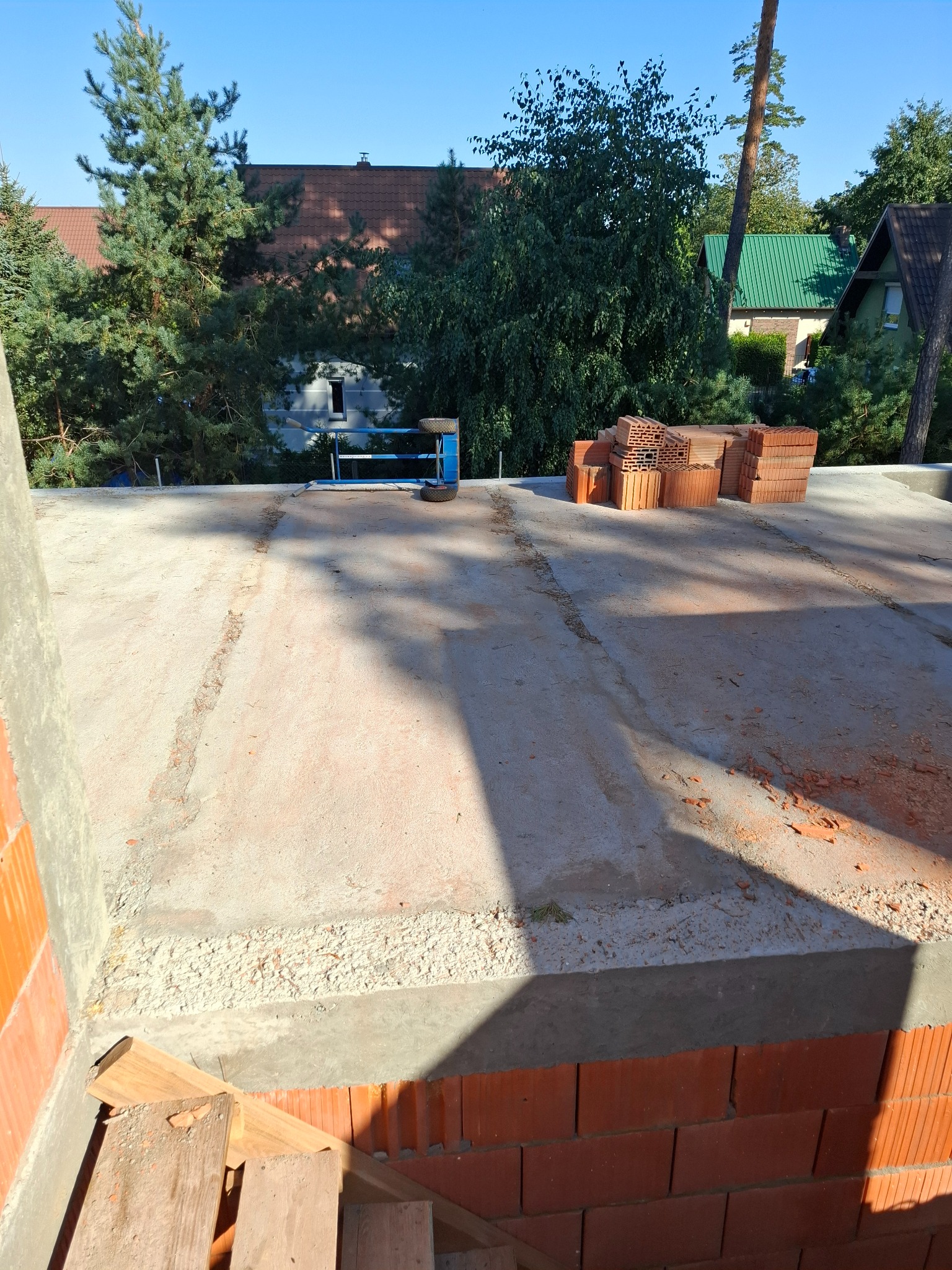 Wylana płyta betonowa z przygotowanymi cegłami na budowie domu jednorodzinnego, widok z góry na tle zieleni i zabudowań.