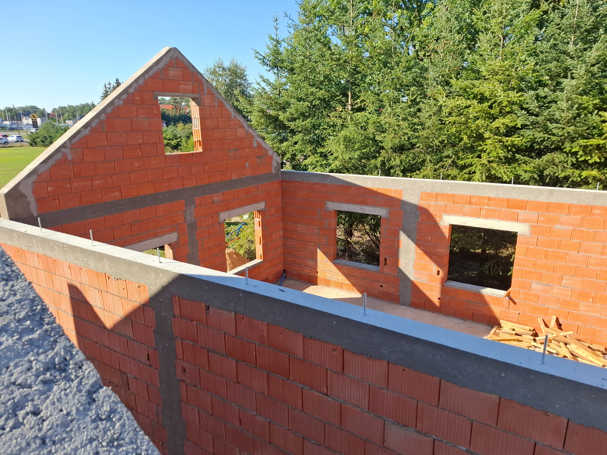 Wznoszenie ścian parteru domu z czerwonej cegły ceramicznej, widoczne otwory okienne i zbrojenie wieńca, w tle zieleń drzew.