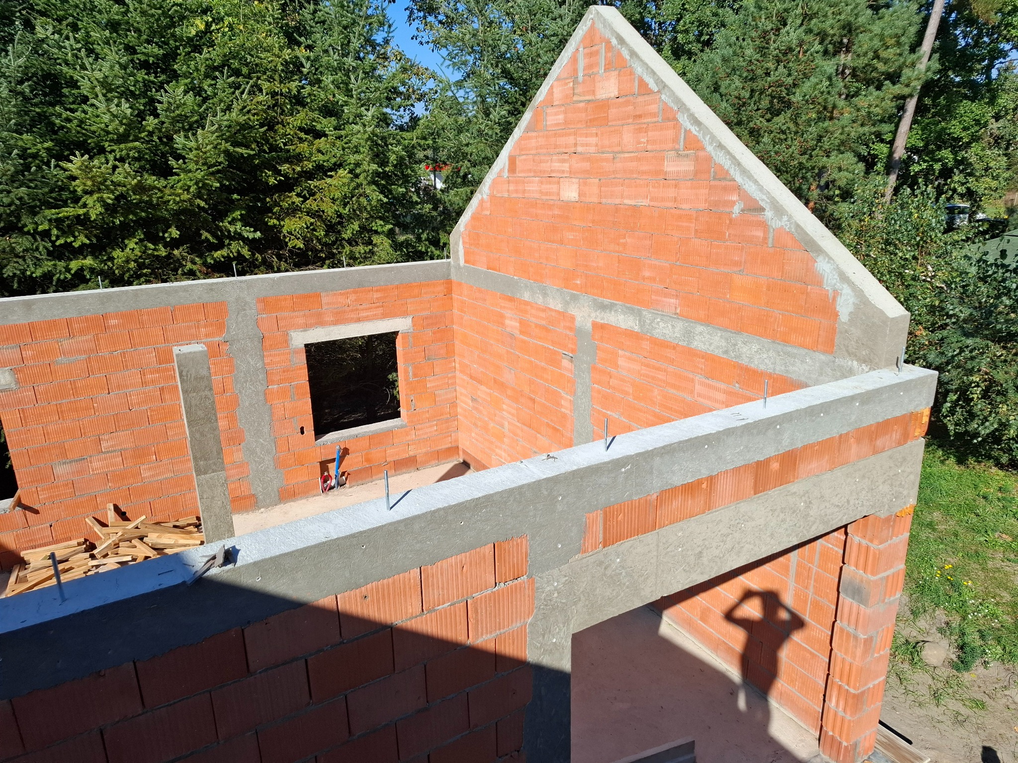 Wznoszenie ścian domu z czerwonej cegły ceramicznej, widoczne zbrojenie i betonowe nadproża, w tle zielone drzewa.