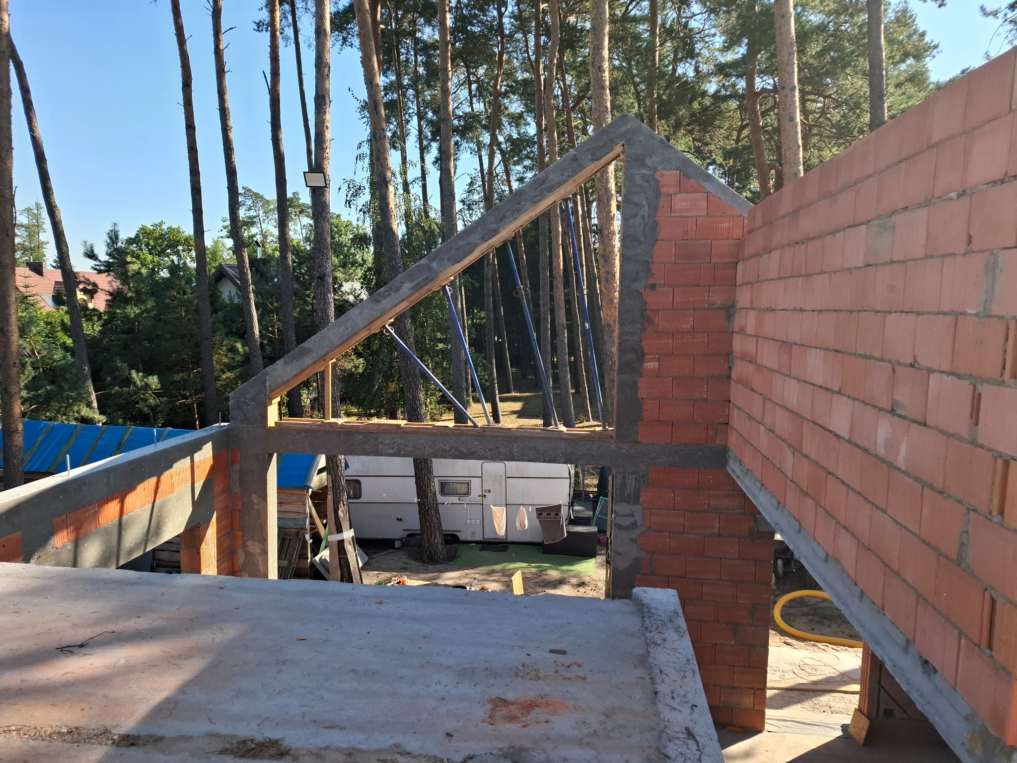 Wznoszenie ścian z czerwonej cegły na budowie domu jednorodzinnego, widoczne szalunki i podpory konstrukcyjne, w tle las i przyczepa kempingowa.