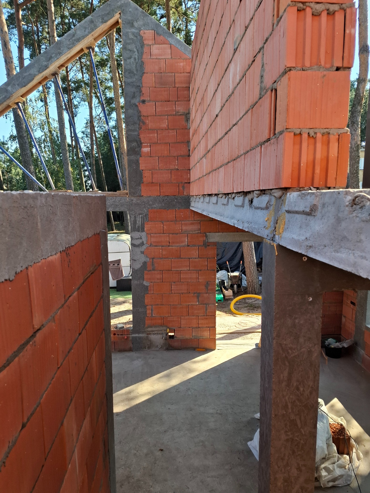 Wznoszenie ścian z czerwonej ceramiki poryzowanej w surowym stanie, widoczne elementy konstrukcji dachu i betonowe nadproża, na zewnątrz drzewa i przyczepa kempingowa.