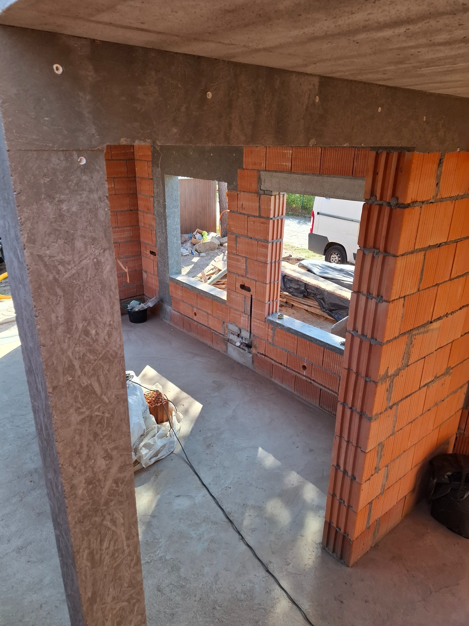 Wnętrze surowego budynku w trakcie budowy, widoczne ściany z czerwonej cegły dziurawki, betonowy strop i otwory okienne, na zewnątrz widoczny fragment białego samochodu dostawczego.