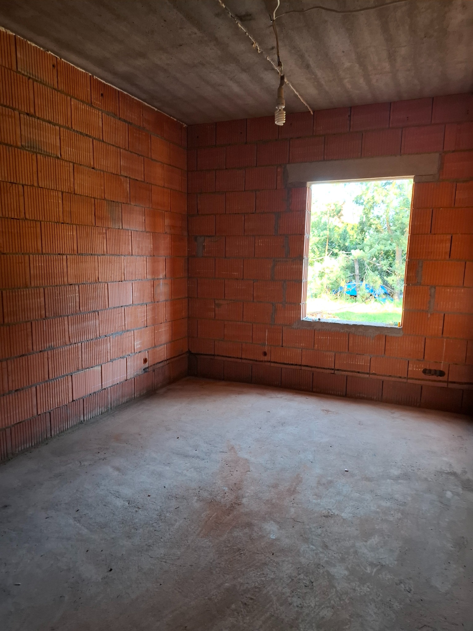 Surowy stan wnętrza domu w trakcie budowy: ściany z czerwonej cegły dziurawki, surowy betonowy strop, okno z widokiem na zieleń, wisząca żarówka.