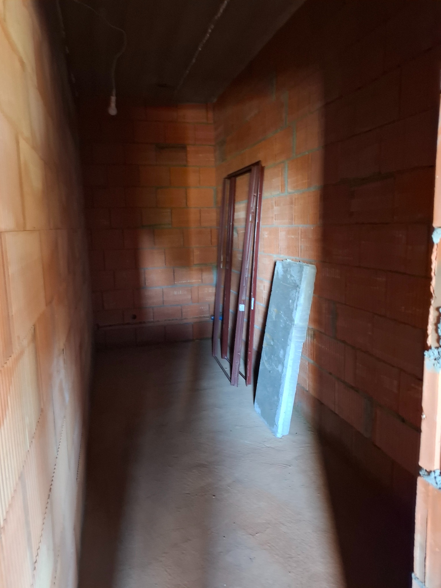 Surowy stan wnętrza z cegły ceramicznej; widoczne ramy okienne oparte o ścianę i betonowy element; żarówka na kablu zwisająca z sufitu.