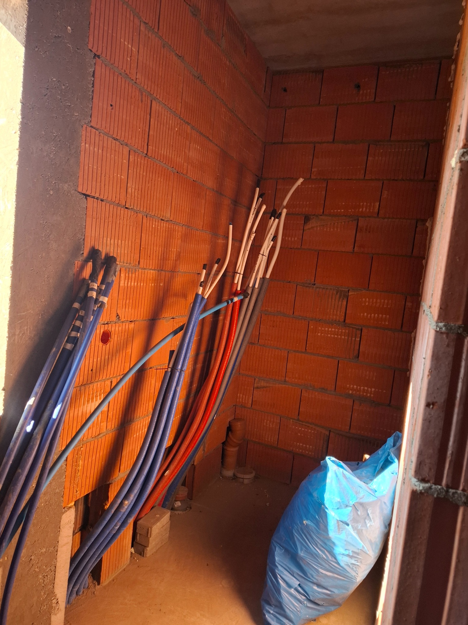 Wnętrze w trakcie budowy: ściany z czerwonej cegły, rury instalacyjne w kolorach niebieskim, czerwonym i białym, oraz niebieski worek na śmieci w rogu.