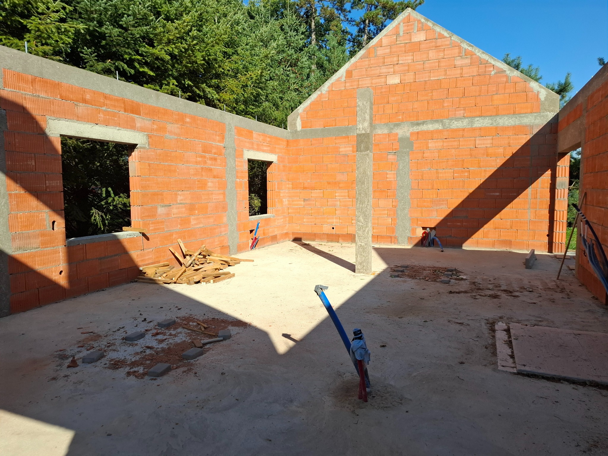 Surowy stan domu z czerwonej cegły, widoczne otwory okienne, betonowe słupy wzmacniające, podłoże betonowe z elementami instalacji, w tle drzewa.