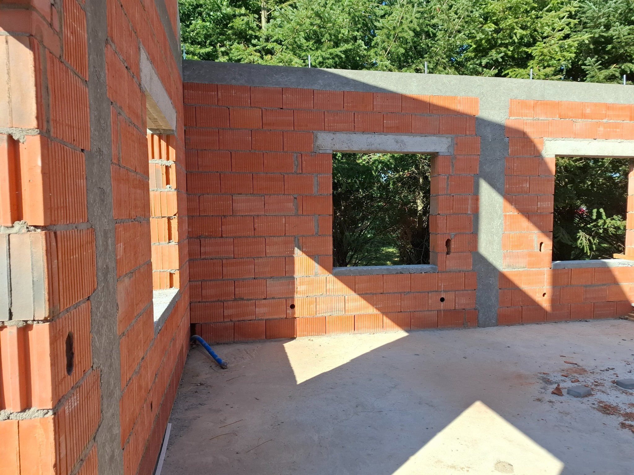 Wnętrze budynku w trakcie budowy, widoczne ściany z czerwonej cegły z otworami okiennymi, częściowo zabetonowane nadproża, niebieski wąż na podłodze, cień od słońca na posadzce.