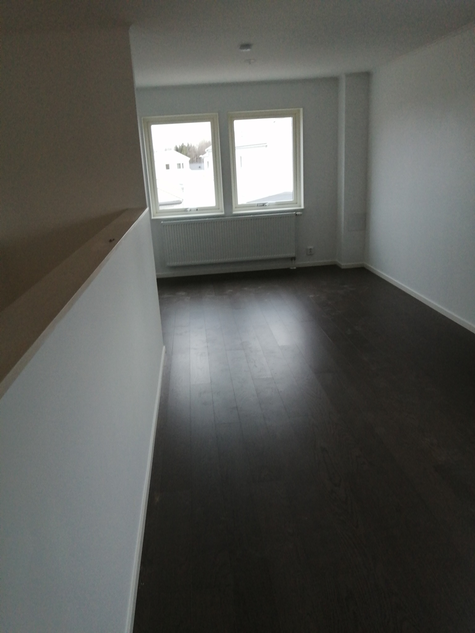 Puste, jasne wnętrze z ciemną podłogą, dwoma oknami z widokiem na zabudowania i grzejnikiem pod oknami, sfotografowane z perspektywy schodów.