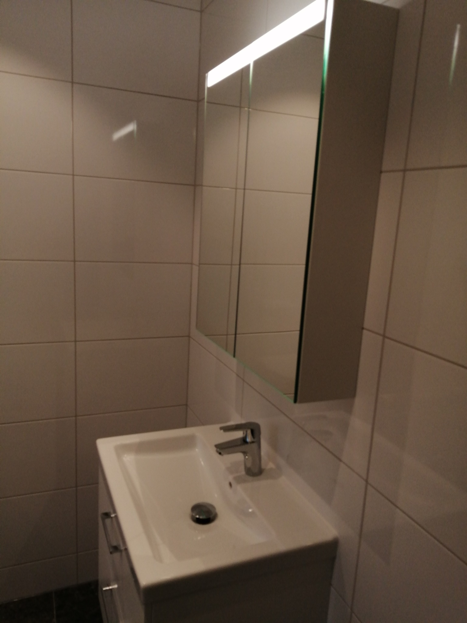 Nowoczesna łazienka w Częstochowie z białymi kwadratowymi płytkami na ścianach, szafką z lustrem z oświetleniem LED nad białą umywalką z chromowaną baterią.