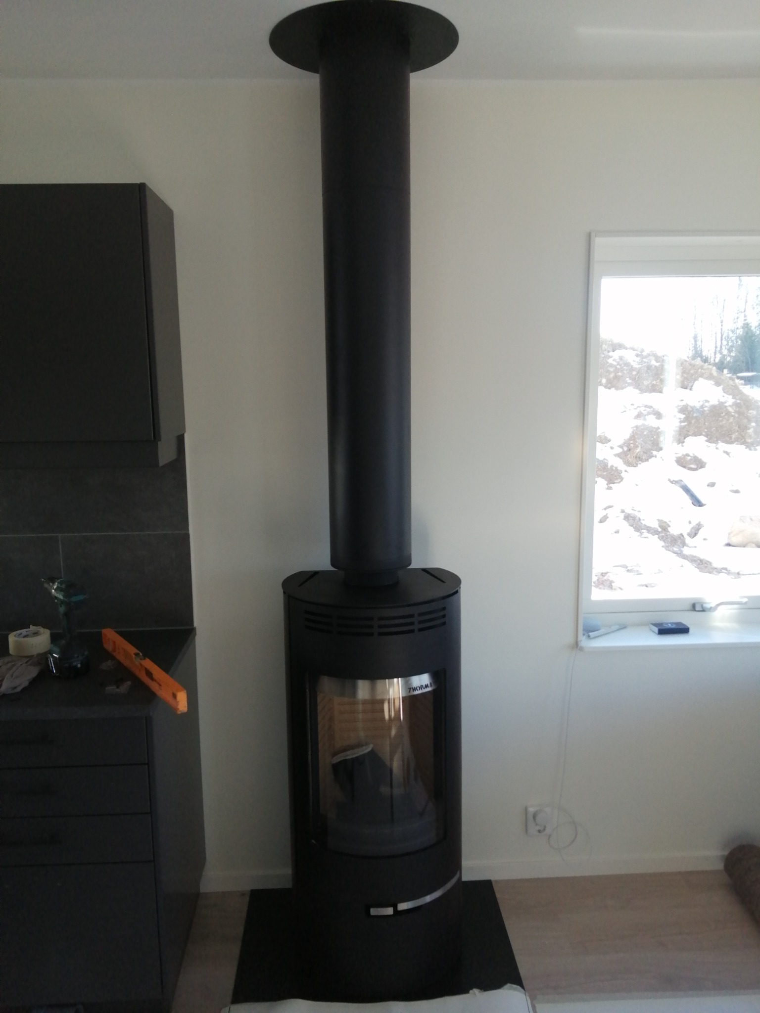 Czarny piec wolnostojący z długim kominem w jasnym wnętrzu, widoczny fragment mebli kuchennych i okno z zimowym krajobrazem.