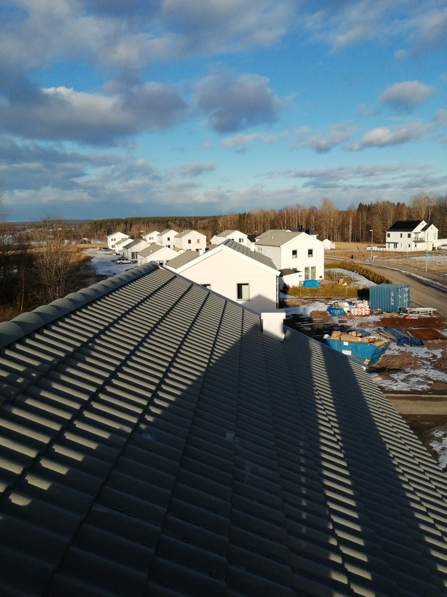 Nowy, szary dach z dachówki ceramicznej w świetle słonecznym, z widokiem na nowo wybudowane domy jednorodzinne i drzewa w tle.
